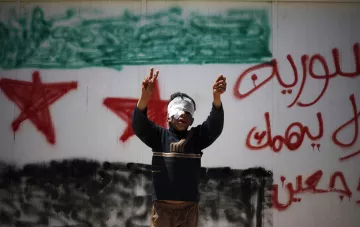 L'image montre un jeune garçon, les yeux bandés, levant les bras en signe de victoire ou de protestation. Il se trouve devant un mur sur lequel sont inscrits des slogans en arabe, ainsi qu'un symbole représentant le drapeau syrien. Cette scène évoque des thèmes de résistance et de luttes dans un contexte de conflit.
