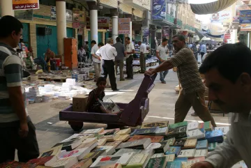 L'image montre un marché animé, probablement un souk, avec des étals de livres disposés au sol. On peut voir plusieurs personnes se déplacer dans la rue, certains feuilletant des livres. Un homme pousse un chariot, où un enfant est assis, observant autour de lui. Les bâtiments en arrière-plan suggèrent une architecture locale, et l'ambiance paraît chaleureuse et vivante.