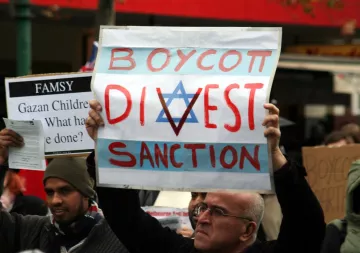 L'image montre des manifestants tenant des pancartes lors d'une manifestation. L'une des pancartes, bien visible, porte les mots "BOYCOTT, DIVEST, SANCTION" avec un symbole en forme d'étoile. Les manifestants semblent exprimés des opinions politiques ou sociales fortes, et l'ambiance de l'événement semble engagée.
