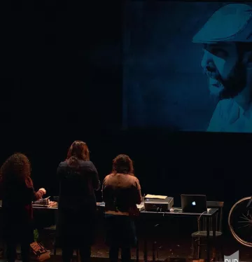 L'image montre un groupe de personnes de dos, se tenant devant une projection qui affiche une image en noir et blanc d'un homme portant un chapeau. Les personnes semblent engagées dans une activité, peut-être en lien avec la projection. Au premier plan, il y a un bureau avec un ordinateur et d'autres objets. L'éclairage crée une atmosphère intrigante autour de la scène.