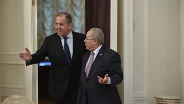 En la imagen se puede ver a dos hombres en un entorno formal, probablemente en una sala de reuniones o un despacho. Uno de ellos, a la izquierda, parece estar señalando o gesticulando con una mano, mientras que el otro, a la derecha, escucha y gira ligeramente su cuerpo hacia él. Ambos visten trajes formales y tienen expresiones serias. El ambiente parece elegante, con detalles en las paredes y una iluminación suave.