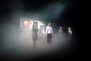 L'image présente une scène de théâtre avec quatre personnages en position debout sur scène. Ils se trouvent dans une atmosphère brumeuse, ce qui ajoute une ambiance mystérieuse. En arrière-plan, on peut apercevoir une petite maison ou un décor qui semble inhabitée. Les personnages sont vêtus de manières diverses, ce qui suggère des rôles ou des traits de caractère différents. La lumière semble tamisée, renforçant l'effet dramatique de la scène.