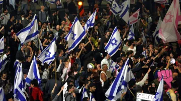 L'immagine mostra una grande folla di persone riunite in un ambiente all'aperto, probabilmente durante una manifestazione. La maggior parte dei partecipanti tiene in mano bandiere di Israele, caratterizzate da una stella di David. Si possono notare anche cartelli, alcuni dei quali contengono frasi e messaggi, mentre altri partecipanti sembrano essere molto energici e coinvolti nell'evento. L'atmosfera appare vibrante e animata, con una forte presenza di colori e simboli nazionali.