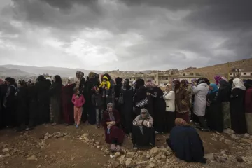 L'image montre un groupe de femmes rassemblées, probablement en attente de quelque chose. Elles portent des vêtements sombres, et certaines tiennent des enfants. Le ciel est nuageux, ce qui donne une ambiance sombre à la scène. En arrière-plan, on aperçoit des maisons sur une colline, soulignant un environnement rural ou de village. L'attente et la solidarité féminine semblent être des thèmes présents dans cette image.