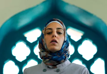 L'image montre une femme portant un hijab coloré, se tenant dans un cadre architectural. La lumière met en valeur son visage et le fond présente des motifs géométriques typiques des designs islamiques, avec des arches et des vitraux. Son expression est sérieuse, ce qui ajoute à la profondeur de la scène.