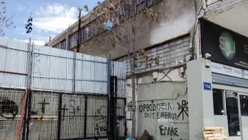 L'image montre un environnement urbain avec un bâtiment en arrière-plan et un mur métallique en façade, où l'on peut voir des graffitis. Un drapeau grec est visible, ainsi qu'un espace de stationnement ou de stockage à côté d'une entrée. Le sol semble en béton, et quelques déchets sont éparpillés sur le sol. Les graffitis sur le mur comprennent des inscriptions en grec, ce qui suggère un contexte local ou culturel. L'atmosphère paraît un peu désordonnée, typique des zones industrielles ou abandonnées.