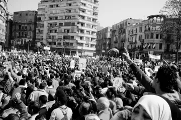 L'image montre une grande foule rassemblée dans une ville, probablement lors d'une manifestation ou d'une protestation. Les participants brandissent des pancartes et semblent exprimer des opinions fortes. L'atmosphère est dynamique et engagée, avec beaucoup de personnes levées les bras, ce qui suggère une ambiance de mobilisation collective. Le noir et blanc de l'image accentue les émotions et l'intensité du moment. Les bâtiments environnants ajoutent un contexte urbain à cette scène de rassemblement.
