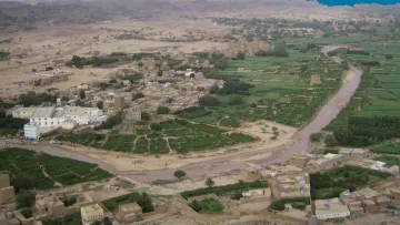 L'image montre un paysage rural, probablement une vallée verdoyante entourée de montagnes arides. Au premier plan, on peut voir des champs cultivés et des terres agricoles, formant un contraste avec les zones désertiques environnantes. Il y a également quelques maisons en terre ou en briques qui semblent faire partie d'un village. La rivière qui serpente à travers le paysage ajoute à la beauté de la scène, symbolisant une source vital d'eau dans cette région dry. Des traces de chemins et des gens peuvent également être aperçus, suggérant une vie active dans cette zone rurale.
