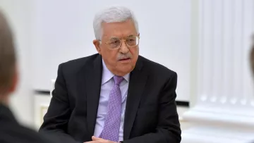 L'image montre un homme assis, portant un costume noir avec une chemise blanche et une cravate mauve. Il a des cheveux gris et porte des lunettes. L'environnement semble formel, probablement une salle de réunion. On peut noter son expression concentrée et attentif pendant la discussion.