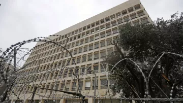 L'image montre un bâtiment imposant avec une architecture moderne, situé sous un ciel nuageux. Au premier plan, des barbelés entourent le périmètre, suggérant une certaine sécurité ou une zone protégée. On peut également apercevoir un arbre, qui contraste avec la structure urbaine. L'atmosphère générale donne une impression de sérieux et de sécurité.