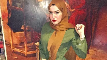 L'immagine mostra una giovane donna che indossa un hijab color marrone e una camicia verde. È in piedi con una mano sulla vita e l'altra sollevata, con un'espressione sicura. Indossa jeans attillati e scarpe marroni. Sullo sfondo si intravede un dipinto o una decorazione murale che aggiunge un tocco di colore all'ambiente. L'illuminazione sembra calda, contribuendo a un'atmosfera accogliente.