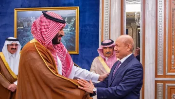 در این تصویر، دو مرد در حال گفت‌وگو و خوش‌آمدگویی به یکدیگر هستند. یکی از آن‌ها لباس سنتی عربی به نام "ثوب" به تن دارد و چفیه‌ای خاص بر سر دارد. او به نظر می‌رسد که در مقام رهبری قرار دارد. مرد دیگر نیز به صورت رسمی لباس پوشیده است و نشان از احترام و ادب در بیان حرکات او دیده می‌شود. در پس‌زمینه، دکوراسیون مجلل و لوکس با تزئینات طلایی و تابلوهایی از مناظر شهری دیده می‌شود که به فضای رسمی این ملاقات افزوده است.