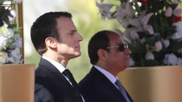L'image montre deux hommes debout, apparemment lors d'un événement officiel. L'un d'eux est en costume sombre avec une cravate, il a une expression concentrée et regarde droit devant lui. L'autre homme, également en costume, porte des lunettes de soleil et semble observateur. Ils se tiennent près de décorations florales, suggérant un cadre cérémonieux.