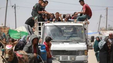 L'image montre un groupe de personnes assises sur le toit d'un camion, tandis que d'autres se trouvent à proximité, y compris des personnes à pied et une charrette tirée par un cheval. L'environnement semble être un endroit animé, avec des tentes et des structures visibles en arrière-plan. On peut voir des fils électriques et des montagnes de déchets, ce qui suggère une atmosphère informelle ou de camp. La scène dégage une impression de communauté et de vie quotidienne dans un contexte peut-être difficile.