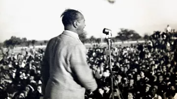 L'image montre un homme en train de parler sur une scène, devant un grand public. Il est vu de dos, et on peut voir une foule nombreuse devant lui, captivée par son discours. On distingue également un microphone sur le podium. L'atmosphère semble dynamique et engagée, probablement lors d'un événement politique ou d'un rassemblement. En arrière-plan, il y a des drapeaux, ce qui pourrait indiquer un contexte symbolique ou patriotique.