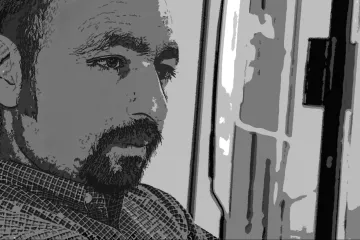 L'image présente un homme dont le visage est partiellement en ombres et lumières, ce qui donne une ambiance introspective. Il semble pensif, regardant dans une direction indéfinie. Les traits de son visage sont marqués, et il a une barbe bien entretenue. Le fond est flou, ce qui permet de concentrer l'attention sur son expression méditative.