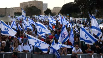 The image shows a large crowd of people gathered together, holding numerous Israeli flags. The atmosphere appears to be part of a demonstration or rally, with many individuals engaged in conversation or focused on the event. The background features buildings and trees, indicating an urban setting. The flags are prominently displayed, creating a sense of unity among the participants.