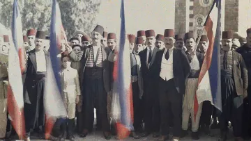 L'image montre un groupe de personnes en tenue traditionnelle, se tenant ensemble avec des drapeaux russes. Les hommes portent des fez et divers vêtements folkloriques, avec un jeune garçon au centre. L'arrière-plan présente une architecture qui semble historique, peut-être un bâtiment symbolique. L'atmosphère suggère un moment de célébration ou de rassemblement associé à une certaine fierté nationale.