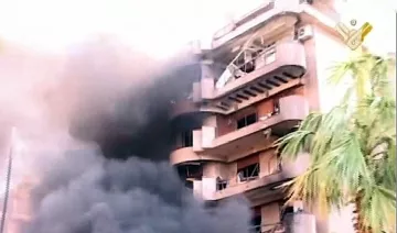 L'image montre un bâtiment en proie à un incendie. De la fumée noire s'échappe des fenêtres, tandis que l'extérieur du bâtiment semble endommagé. Des éléments de végétation, comme un palmier, sont visibles au premier plan, contrastant avec la scène chaotique et la destruction. Le ton général de l'image est sombre, mettant en évidence l'urgence et la gravité de la situation.
