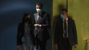L'image montre trois personnes dans un hall. Deux d'entre elles portent des masques, et on peut voir qu'elles sont habillées de manière formelle. La personne à gauche semble avoir un badge autour du cou. Les trois individus se tiennent près d'un mur, avec une lumière dorée en arrière-plan. L'ambiance semble professionnelle et sérieuse.
