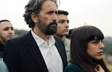 L'image montre un groupe de personnes rassemblées, avec un homme au premier plan, portant un costume noir et une chemise blanche. Il a les cheveux gris et une barbe. À côté de lui se trouve une femme avec des cheveux longs et bruns, qui semble pensive. En arrière-plan, d'autres personnes sont présentes, affichant des expressions sérieuses. L'ambiance général semble solennelle.