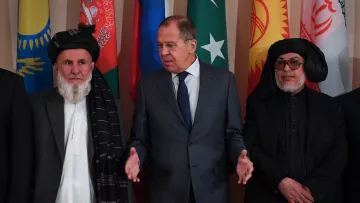 The image features a group of men standing in front of a backdrop adorned with various national flags. In the center is a man in a suit, possibly a diplomat or official, gesturing as he speaks. To his left and right are two men dressed in traditional attire, including a shalwar kameez and a shawl. The overall atmosphere suggests a formal meeting or diplomatic discussion, likely concerning regional issues or cooperation.