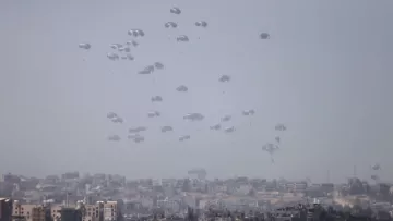 L'immagine mostra un cielo grigio-azzurro con numerosi paracadute che scendono lentamente verso un'area urbana sottostante. Sullo sfondo, è visibile una città con edifici di varie dimensioni e forme. L'atmosfera sembra tesa, suggerendo che si tratta di un'operazione militare o di emergenza. I paracadute, bianchi e leggeri, creano un contrasto visivo con l'ambiente circostante.
