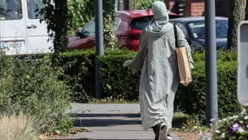 L'image montre une personne marchant sur un chemin. Elle porte une longue robe de couleur claire et un foulard. Elle tient un grand sac en papier dans une main. Le décor environnant inclut des buissons et des arbres, avec des voitures en arrière-plan. La scène semble tranquille et ensoleillée.