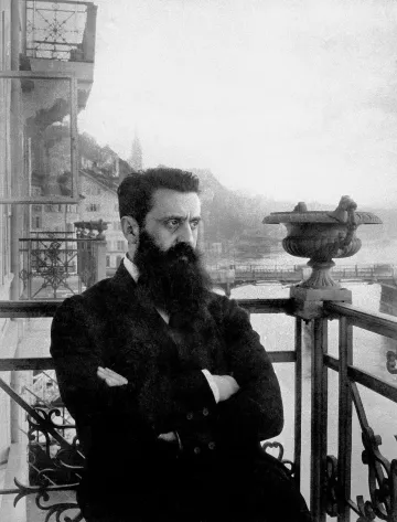 L'image montre un homme avec une barbe fournie, portant un costume sombre. Il est debout sur un balcon orné de balustrades métalliques, avec un regard pensif et sérieux. En arrière-plan, on aperçoit des montagnes et un plan d'eau, suggérant un paysage pittoresque. L'atmosphère est nostalgique, renforcée par le ton en noir et blanc de la photographie, ajoutant une dimension historique à la scène.