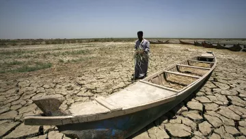The image depicts a man standing on cracked, dry land beside a wooden boat. The landscape appears parched, indicating a lack of water, possibly due to drought. The man is holding a bundle of vegetation or grass. In the background, other boats are visible, also resting on the dry ground. The scene conveys a sense of environmental distress and the challenges faced by individuals in regions affected by water scarcity. The sky is clear, and the sunlight emphasizes the harsh conditions of the landscape.