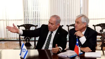 The image shows two men engaged in discussion at a table. One man is gesturing with his hand while speaking, suggesting he is making a point or explaining something. He is wearing a suit and a tie with stripes. The other man is sitting close by, listening attentively. He has gray hair and is also dressed in a suit. In front of them, there are flags of Israel and Chile on the table, indicating an official meeting or diplomatic discussion. The background features large windows with blinds, allowing natural light into the space.