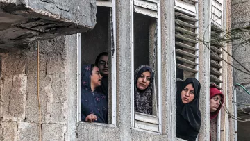 L'image montre plusieurs femmes regardant par des fenêtres d'un bâtiment. Elles semblent attentives et curieuses, certaines portant des hijabs. Le mur extérieur du bâtiment est en béton, et on peut apercevoir des fenêtres aux volets cassés. L'ambiance paraît chargée d'émotions, mais on ne peut pas déterminer le contexte exact de la scène.