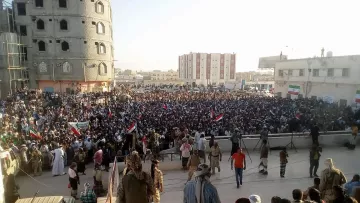 L'image montre une grande foule rassemblée lors d'une manifestation ou d'un rassemblement public. Les participants tiennent des banderoles et des drapeaux, indiquant un fort engagement politique ou social. En arrière-plan, on peut voir des bâtiments en construction ou rénovés, ainsi qu'une architecture variée. La scène semble se dérouler en plein air, probablement durant l'après-midi ou le début de la soirée, avec une lumière naturelle éclatante. L'atmosphère est dynamique, avec beaucoup de gens engagés dans une cause commune.