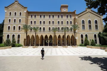 L'image montre un grand bâtiment en pierre avec une architecture traditionnelle, comportant plusieurs fenêtres à arc et des colonnes. Au premier plan, une personne marche seule sur une place pavée, tandis que des palmiers sont visibles sur les côtés. Le ciel est clair et bleu, ce qui donne une atmosphère ensoleillée. Le bâtiment semble être une institution éducative ou un lieu historique.