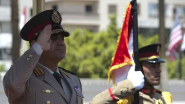 L'image représente deux officiers militaires en uniforme. L'un d'eux, probablement un haut responsable, effectue un salut militaire, tandis que l'autre maintient un drapeau. L'environnement semble être une cérémonie officielle en extérieur, avec des bâtiments en arrière-plan. Les uniformes sont ornés de médailles et de distinctions, reflétant un certain niveau de cérémonie et de formalité.