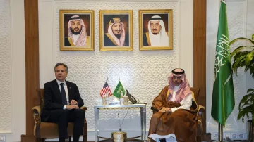 در تصویر، دو نفر در یک اتاق جلسه نشسته‌اند. در سمت چپ، یک مرد با کت و کراوات سیاه قرار دارد که به نظر می‌رسد فردی رسمی باشد. در سمت راست، مردی با لباس سنتی عربی نشسته است. پشت آن‌ها سه پرتره در قاب زرد رنگ قرار دارد که به نظر می‌رسد تصاویری از شخصیت‌های مهم هستند. همچنین، پرچم‌های ایالات متحده و عربستان سعودی روی میز قرار دارند و یک پرچم دیگر عربستان نیز در کنار آن‌ها دیده می‌شود. محیط اتاق با تزئینات مدرن و گیاهان تزئینی آراسته شده است.