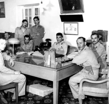 L'image montre un groupe de six hommes en uniforme militaire réunis autour d'une grande table en bois dans un bureau. Certains d'entre eux sont assis, tandis que d'autres se tiennent derrière. L'environnement décoré de manière classique inclut des éléments tels qu'une lampe de bureau et des tableaux accrochés au mur. L'atmosphère semble formelle, et les expressions des hommes varient entre concentration et sérieux.