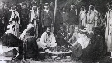 L'image montre un groupe de personnes, probablement issus de cultures arabes, rassemblées autour d'un grand plat de nourriture posé au sol. Certains sont assis tandis que d'autres se tiennent debout en arrière-plan. Les participants portent des vêtements traditionnels, tels que des robes et des turbans, ce qui suggère un contexte historique. L'ambiance semble conviviale, et l'événement pourrait être un repas ou une célébration. Des éléments du décor indiquent que la scène se déroule en extérieur, possiblement sous une tente.
