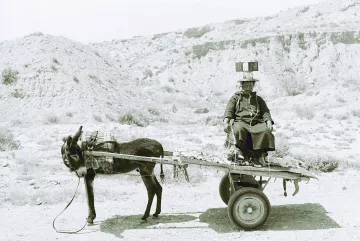 L'image montre une scène en noir et blanc d'un homme assis sur un chariot tiré par un âne. L'homme porte des vêtements traditionnels et semble se diriger quelque part, tandis que l'âne est attaché à l'avant du chariot. L'arrière-plan présente un paysage aride et désertique, avec des collines ou des montagnes à l'horizon. L'atmosphère de la photographie évoque un sentiment de simplicité et de vie rurale.