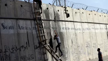 L'immagine mostra due persone che stanno scalando un muro di cemento, sul quale è presente della rete metallica. Dietro di loro, è visibile una scala che qualcuno sta utilizzando per salire. Il muro è coperto da graffiti e scritte, in varie lingue, che sembrano affrontare temi legati alla storia e alla condizione sociale. La scena evoca un senso di tensione e disperazione, con le persone che tentano di superare un ostacolo fisico, simbolo di divisione.