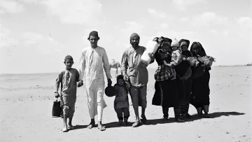 L'image montre un groupe de personnes marchant sur une surface sablonneuse, probablement dans un désert. Au premier plan, on peut voir plusieurs enfants et adultes. Les adultes portent des vêtements traditionnels. Certains membres du groupe transportent des objets sur leurs épaules ou dans les bras, tandis que les enfants semblent tenir la main de l'un des adultes. L'arrière-plan est vaste et dégagé, avec quelques nuages dans le ciel. L'atmosphère dégage une impression de déplacement ou de voyage.