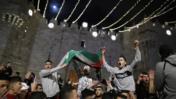 L'image montre un rassemblement nocturne devant un bâtiment ancien, probablement une fortification. Des personnes sont réunies en célébrant, certaines agitant un drapeau palestinien. L'atmosphère semble festive et dynamique, avec des lumières scintillantes qui illuminent la scène. Les expressions des participants retranscrivent une ambiance d'excitation et d'engagement.