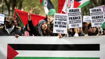 تظهر الصورة مجموعة من المتظاهرين في شارع، يحملون لافتات وشعارات تتعلق بفلسطين. يوجد علم فلسطيني في مقدمة المظاهرة، ويتوسط الحشد شخص يرفع ذراعيه بعلامة النصر. اللافتات تحمل عبارات تدعو لدعم فلسطين وتندد بفعل الإسراء وتتهم فرنسا بالتواطؤ. المشهد يعكس شعورًا قويًا بالتضامن مع القضية الفلسطينية.