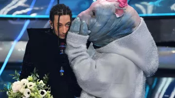 L'image montre une scène d'un spectacle où une personne au style vestimentaire unique, tenant un bouquet de fleurs, semble être en conversation avec une autre personne déguisée dans un costume original. Le costume ressemble à un animal aquatique, avec une tête d'apparence bleue et des détails colorés. L'ambiance semble dramatique ou émotive, avec un éclairage qui accentue les éléments de la scène.