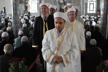 L'image montre un groupe d'hommes portant des vêtements traditionnels, notamment des robes blanches et des turbans. Ils semblent être en procession, probablement lors d'un événement religieux. En arrière-plan, de nombreuses personnes sont assises, probablement en prière ou en attente d'une cérémonie. L'architecture de la pièce inclut des motifs décoratifs et des fenêtres qui laissent entrer la lumière, créant une atmosphère solennelle.