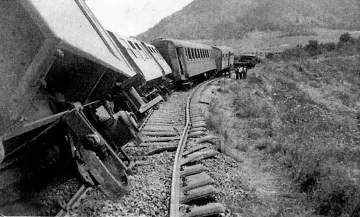 L'image montre un accident de train. Plusieurs wagons sont déraillés, penchés sur le côté, tandis que les rails semblent déformés. En arrière-plan, on aperçoit des montagnes et des personnes qui semblent inspecter les lieux. L'atmosphère est celle d'un événement tragique, évoquant une situation d'urgence.