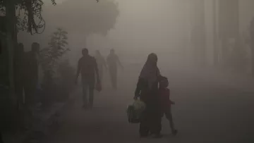 L'image montre une scène urbaine plongée dans une atmosphère brumeuse ou poussiéreuse. Dans le premier plan, une femme portant un hijab tient la main d'un jeune garçon. Ils semblent marcher ensemble le long de la rue. En arrière-plan, d'autres silhouettes de personnes sont visibles, floues par la brume, tandis que des arbres et des structures urbaines apparaissent à peine à travers la poussière. L'ambiance semble calme mais chargée d'un certain mystère à cause de la visibilité réduite.