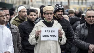 L'image montre un groupe de personnes rassemblées, dont certaines portent des vêtements d'hiver. Au premier plan, un homme se distingue en tenant une pancarte sur laquelle est écrit « PAS EN MON NOM ». Son expression est sérieuse et déterminée. Les autres personnes autour de lui semblent également concentrées sur la situation. L'ambiance générale de la scène suggère une manifestation ou un rassemblement pour une cause particulière.
