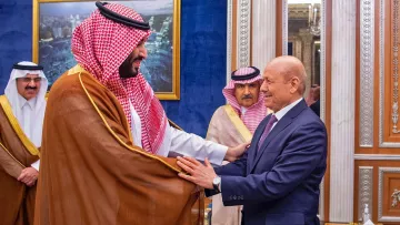 L'image montre deux hommes en costume, engagés dans une interaction amicale, probablement lors d'une rencontre officielle. L'un des hommes porte un habit traditionnel avec un châle. L'arrière-plan présente un décor élégant, typique d'un palais, avec des éléments architecturaux raffinés. D'autres personnes en tenue formelle sont visibles, suggérant un contexte diplomatique ou cérémoniel.