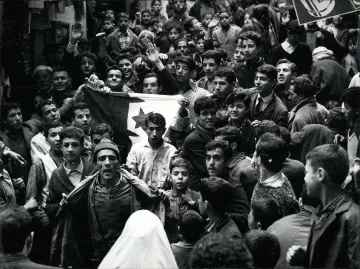 L'image montre une foule dense de personnes rassemblées dans une rue. Au premier plan, plusieurs individus sont visibles, dont un homme qui semble porter un drapeau avec un symbole distinctif. Les expressions des visages varient, certains levant les bras en signe de soutien ou d'enthousiasme. On peut observer des jeunes, des adultes et même des enfants parmi la foule. L'atmosphère semble intense et chargée d'émotion, indiquant un moment significatif ou une manifestation. La scène est en noir et blanc, ce qui lui donne un aspect historique.